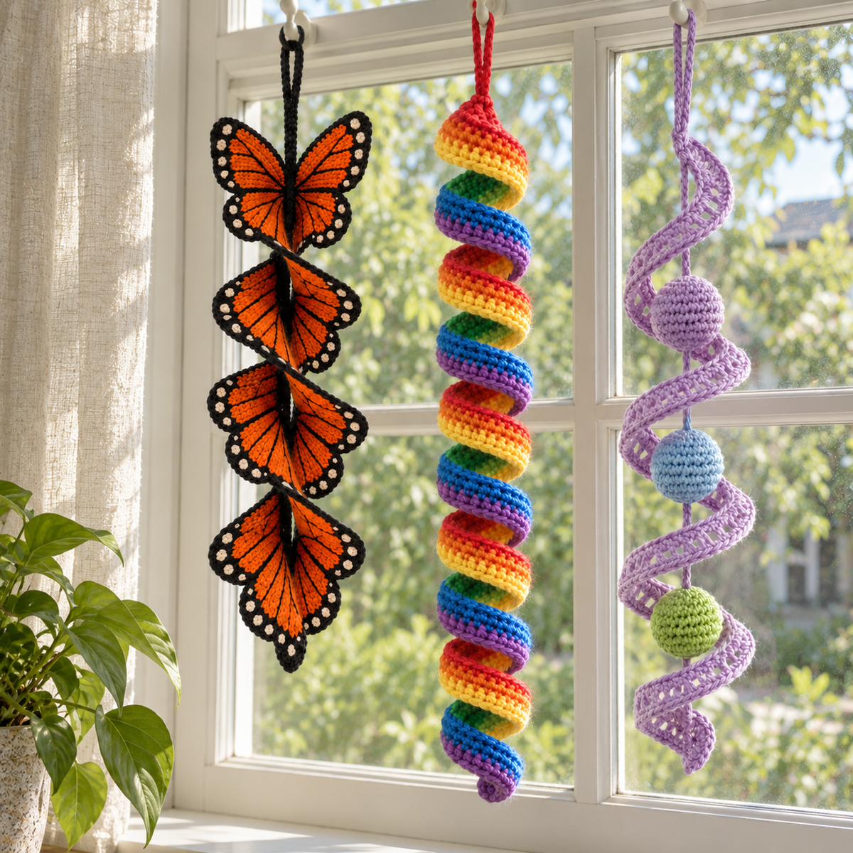 An orange and black Monarch butterfly spinner, a rainbow ribbon spiral and a lilac lace twist with three coloured balls hanging side by side in a sunny window.