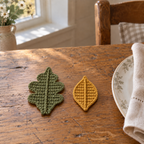 An olive lobed oak leaf and a mustard oval leaf with raised centre ridges beside a linen napkin and plate.