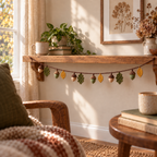 Six acorns and six olive or mustard crochet leaves on a brown garland hanging beneath an oak shelf in a cosy reading corner.