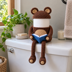 Brown Bear reading roll holder with tan-lined round ears and an open blue book, sitting on the front of a white toilet cistern.