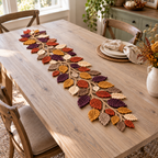 A dense crochet leaf runner in plum, rust, mustard, ivory and taupe on tan branches across a pale wood dining table.