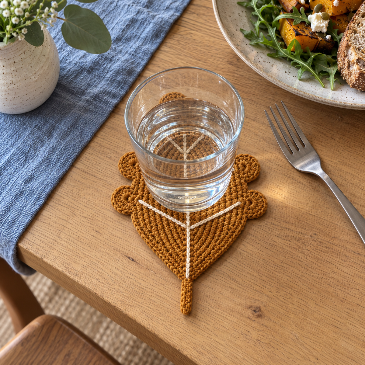 A clear water glass on an ochre Golden Oak crochet leaf coaster with rounded lobes and cream veins on a wooden lunch table beside blue linen and a fork.