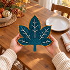 Two adult hands supporting a finished Teal & Gold crochet leaf mat above a wooden table beside a ceramic plate.