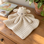 Cream lamb crochet lovey with a bobble crown and softly gathered blanket on a dresser beside two picture books and a leafy plant.
