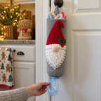 Heather-grey gnome bag holder with a red front hat, white looped beard and pink nose hangs on a pantry knob while a hand pulls a blue plastic shopping-bag handle from the bottom.