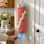 A hand pulls a shiny light-blue shopping bag handle first from the bottom of a coral crochet holder with seven white daisies, while coloured bag handles show at its open top.