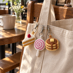 A miniature layered cake slice, swirl lollipop and three-pancake stack hanging from a canvas tote in a bright cafe.