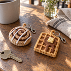 A close view of a miniature coiled cinnamon roll with white icing and a square grid-textured waffle with butter and syrup, both crocheted in matte cotton.