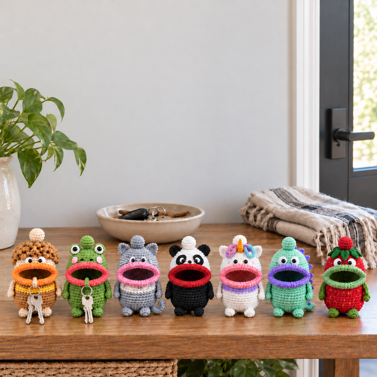 Crochet key pouches Curly Mop, Frog, Cat, Panda, Unicorn, Dino, Strawberry stand on a wooden entryway console beside a key bowl and scarf.