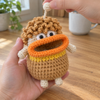 The same Curly Mop key pouch is held in a kitchen while the other hand lifts its cream stopper ball above the crown. The keys are tucked inside its open mouth.