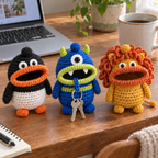 Penguin, a one-eyed blue Monster and Lion crochet key pouches stand on a wooden desk beside a laptop and mug. Two house keys hang from the Monster mouth.