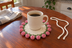 Rosebud: a coffee mug on a flat warm-white crochet coaster edged with rose-pink popcorn buds and green leaves.