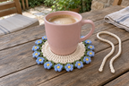 Forget-me-not: a pink coffee mug on a flat sky-blue flower crochet coaster with yellow centres and green leaves.