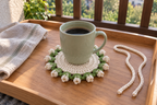 Lily of the Valley: a coffee mug on a flat warm-white crochet coaster edged with ivory bells and green leaves on a breakfast tray.