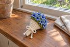 Cornflower: a warm-white crochet bouquet with pointed blue cornflowers, dark centres and a white bow on a windowsill.