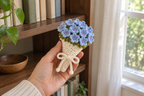 Hydrangea: a hand holding a tied warm-white crochet bouquet with blue hydrangea florets and pale-lilac centres.