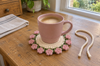 Peony: a pink coffee mug on a flat warm-white crochet coaster edged with full blush-pink peonies and green leaves.