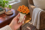 Marigold: a hand holding a tied warm-white crochet bouquet with frilled orange marigolds and darker centres.