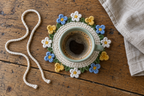 Mixed Wildflower: a green coffee cup on a flat warm-white crochet coaster with alternating white, gold and blue flowers and a loose white tie cord.