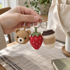An adult hand holding a crochet bear head, strawberry and coffee cup charm by their rings.