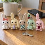 Five crochet bunny keychains with bow, egg, scarf, cherries and heart details beside a mug and phone on a desk.