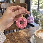 An adult hand holding a donut and a macaron crochet charm by their rings.