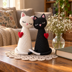 White and black crochet cats with red hearts and crossing tails on a coffee-table doily beside a book and a vase of delicate white flowers.