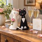 White bridal and black tuxedo crochet cats with a mesh veil, flower crown and three-rose bouquet beside two champagne glasses and a floral wedding card.