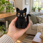 An adult hand holds a small finished black crochet cat upright, showing its amber stitched eyes, pointed ears, front paws and curved tail in a living room.