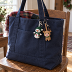Snowman and reindeer crochet keychains hang from the handle of a navy winter tote bag on a wooden chair.