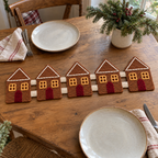 Five caramel gingerbread houses with cream roof trim, gold windows and burgundy doors joined as a crochet runner on a wooden table between two place settings.