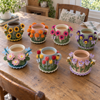 Seven Flower Garden crochet coasters hugging plain mugs on a wooden table: Sunflower Patch, Marigold Glow, Gerbera Bright, Pansy Faces, Water Lily Pond, Tulip Row, Crocus Awakening.