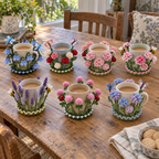 Seven Flower Garden crochet coasters hugging plain mugs on a wooden table: Bluebell Wood, Rose Garden, Peony Blush, Carnation Posy, Lavender Field, Clover and Ladybirds, Hydrangea Cloud.