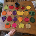 Fruit crochet coasters on a table: Pear, Red Apple, Strawberry, Cherry, Lemon, Orange, Peach, Plum, Banana, Pineapple, Whole Watermelon, Grape Bunch, Pomegranate, Fig, Mango, Raspberry, Whole Avocado, Persimmon, Dragon Fruit, Starfruit, Cantaloupe Melon.