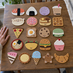 Kawaii Food crochet coasters on a table: Salmon Nigiri, Onigiri, Mochi, Bubble Tea, Dumpling, Iced Donut, Pancake Stack, Square Waffle, Pizza Slice, Egg Toast, Chocolate Chip Cookie, Macaron, Ramen Bowl, Taco, Burger, Cupcake, Cake Slice, Fluted Tart, Ice Cream Cone, Gingerbread Star Cookie, Croissant.
