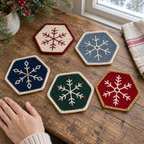 Five hexagonal Christmas Snowflake crochet coasters on a wooden table beside a hand: Heritage Branch, Frosted Star, Midnight Crystal, Evergreen Sprig, Ruby Lace.