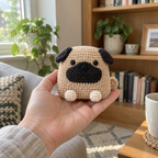 An adult hand holding a small fawn crochet Pug upright in a living room, showing its dark ears and muzzle, cream paws and curled side tail.