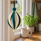 A teal and ivory Classic Droplet hangs from a wooden peg beside a kitchen window, with a ceramic mug and herb pot on the sill.