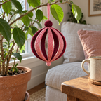 A rose and burgundy Round Bauble hangs from a houseplant branch beside a window and sofa, near a wooden side table and mug.