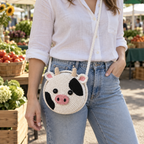 A white round Cow crochet crossbody bag with black patches and a pink muzzle worn at the hip by an adult in a white shirt and denim jeans at a farmers market.