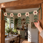 An adult hand gently supports a sage leaf branch of Botanical Bloom while the finished cream-and-sage flower hanging remains tied across a wooden kitchen doorway.