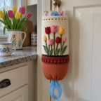 A cream crochet bag holder with upright red, pink and yellow tulips, sage leaves and a terracotta pot hangs from a pantry knob, with a blue plastic shopping-bag handle at the bottom.