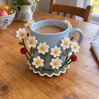 White crocheted daisies and two red ladybirds surrounding a pale-blue mug of tea on a sage-green crochet coaster.