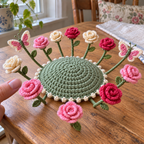 A fingertip beside pink, deep-red and cream crochet roses surrounding the clear raised centre of a sage-green coaster with two butterflies.