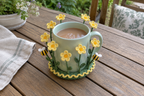 Yellow crochet buttercups and two bees surround a pale-green mug of tea on a wooden patio table.