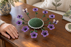 Violet crochet anemones and two butterflies surround the clear raised centre of a deep-green coaster beside a resting hand on a wooden sideboard.