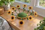 Golden crochet sunflowers with brown centres and two bees fan outward from a leaf-green domed coaster with golden edging on a bamboo window ledge.