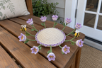 Lilac-and-white crochet crocus cups and two bees fan outward from a cream domed coaster with lilac edging on a wooden table.