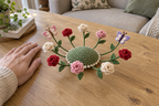 Red, pink and cream crochet roses and two butterflies fan outward from a sage-green domed coaster beside a resting hand on a wooden table.