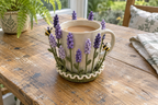 Lavender and violet crochet flower spikes and two bees surround a cream mug of tea on a wooden sunroom table.